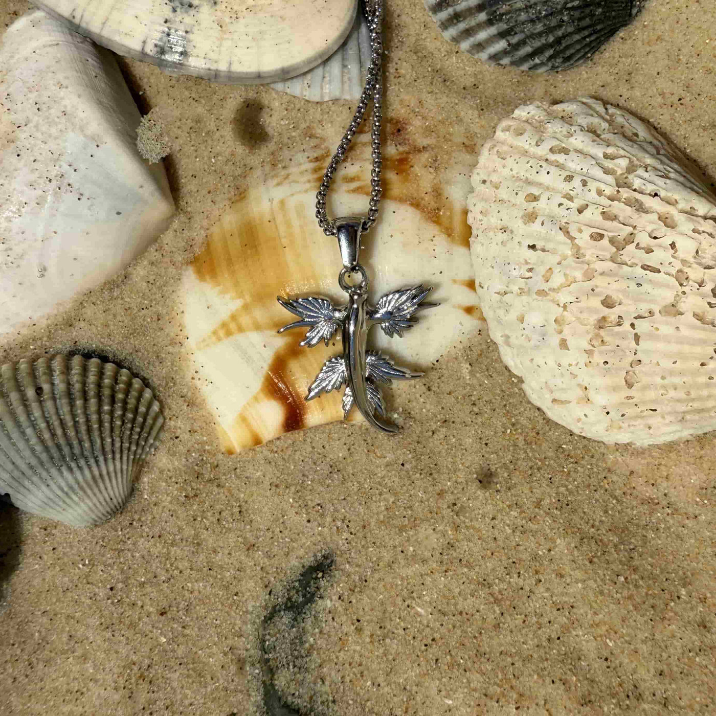 A silver-toned metal pendant necklace with a stylized winged cross hangs on a chain and rests on sand surrounded by seashells.